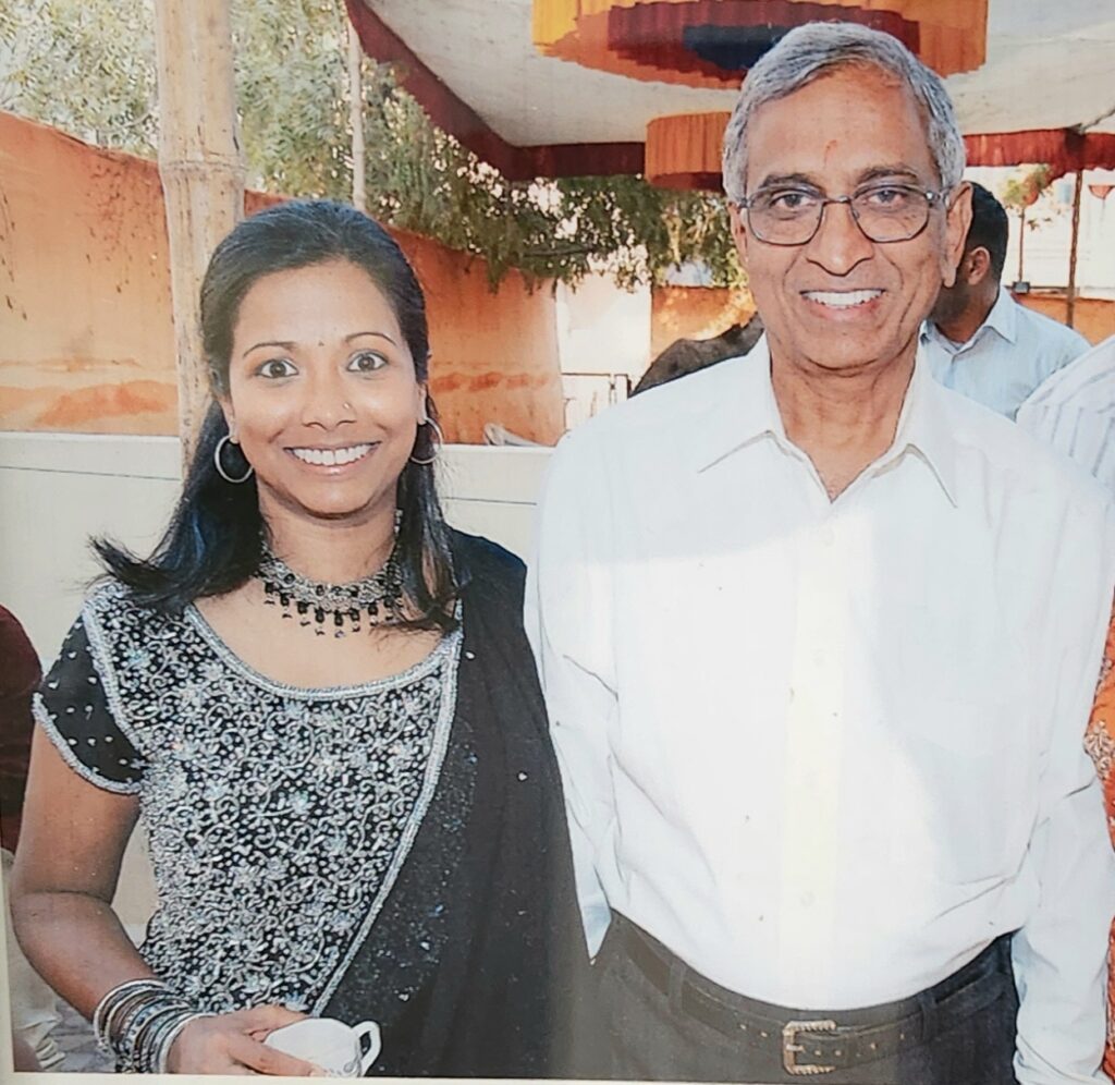 A smiling Indian woman wearing a black and white sari next to an older Indian man in a white button-up. 